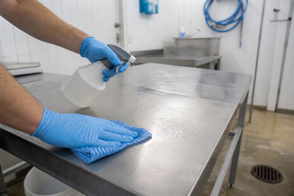 Gloved hands sanitising a stainless steel preparation surface in a bright, orderly space